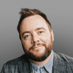 Professional headshot of a smiling man with a beard and short dark hair, wearing a grey plaid blazer over a blue collared shirt.