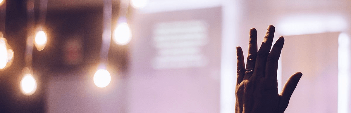 Person's hand raised at an event, with blurred atmospheric lights and a projected screen in the background.